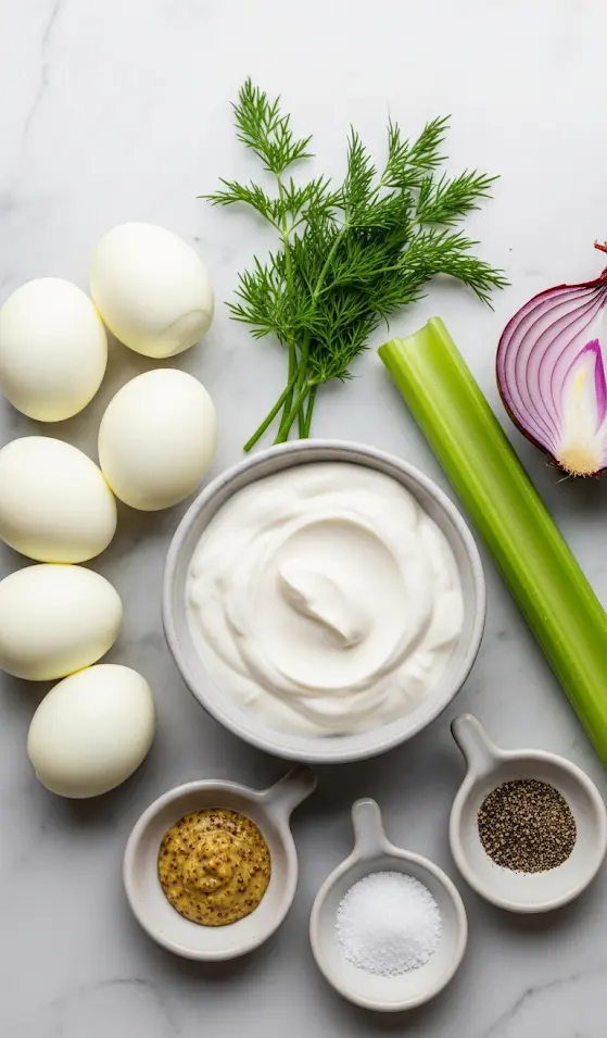 A flat lay of fresh ingredients for egg salad: eggs, Greek yogurt, celery, red onion, dill, and mustard on a marble surface.